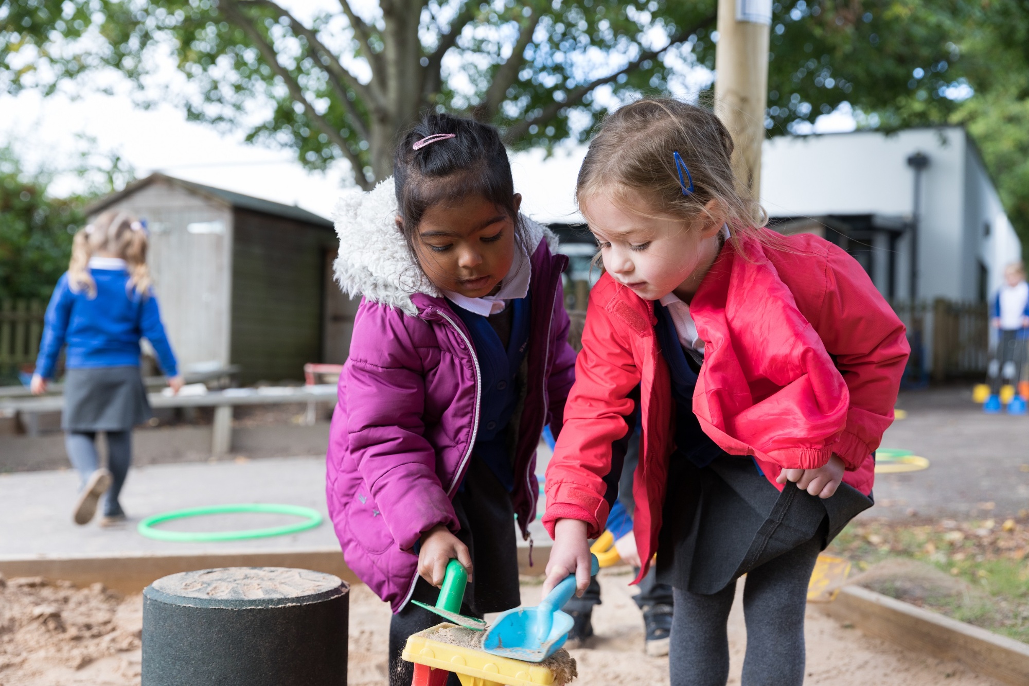 Two children tap a bucket with spades in the sand pit